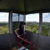 Wide angle image of women in fire tower with thousands of acres of trees in distance.