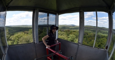 Wide angle image of women in fire tower with thousands of acres of trees in distance.